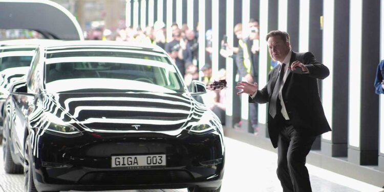 Elon Musk gesturing near a black Tesla vehicle in a modern setting.